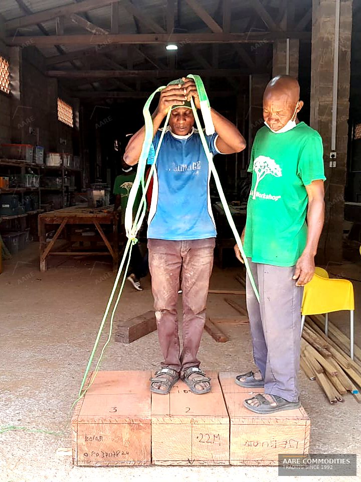 Two AARE Commodities yard hands preparing lifting slings while standing on squared Ekki timber baulks inside a Nigerian timber workshop