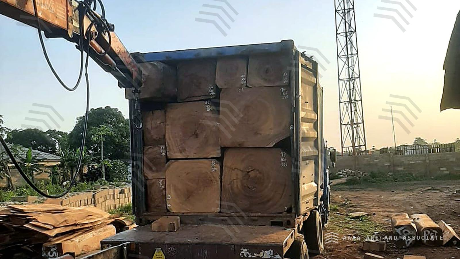 Shipping container on a truck packed with squared hardwood baulks, each end marked with dimensions, alongside a truck-mounted crane at dusk