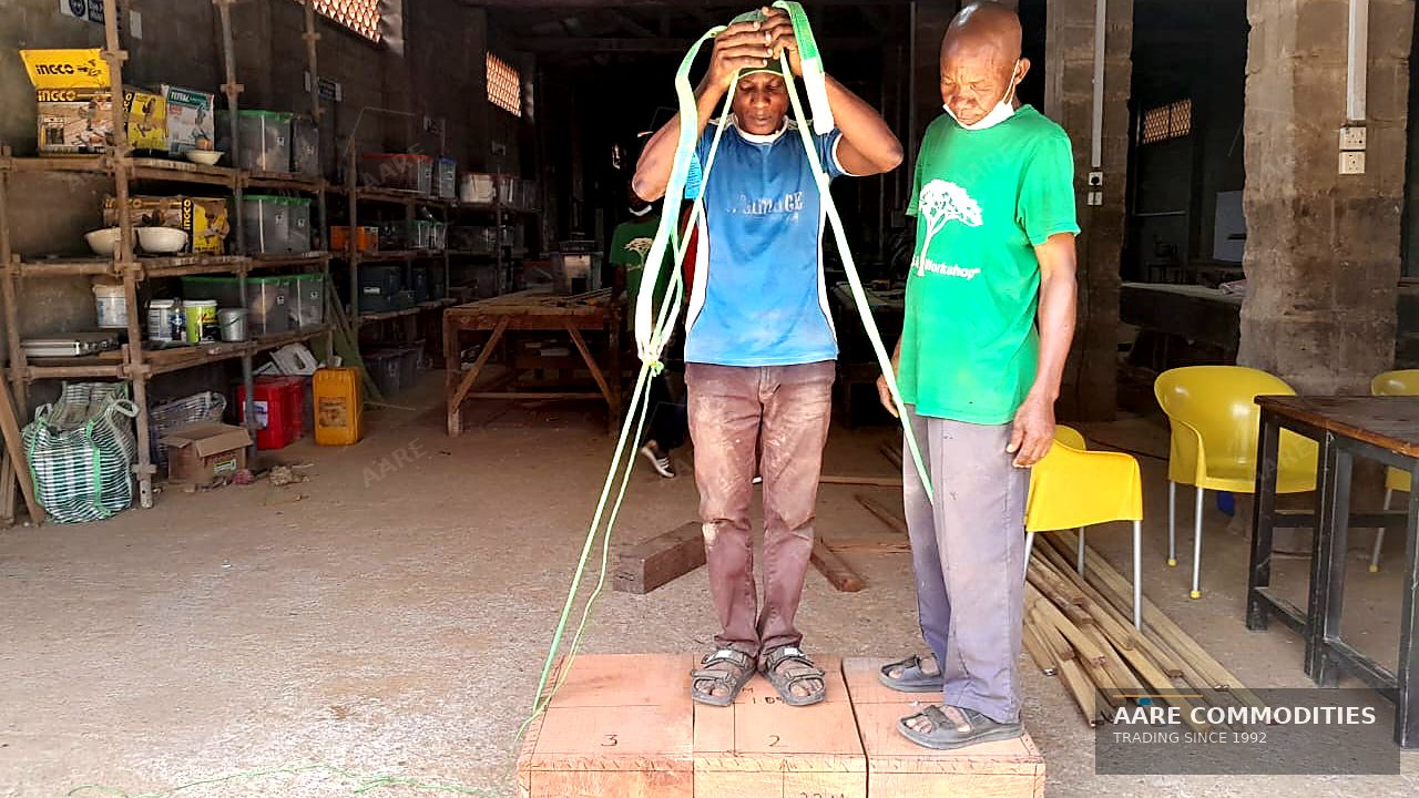 Two AARE Commodities yard hands preparing lifting slings while standing on squared Ekki timber baulks inside a Nigerian timber workshop